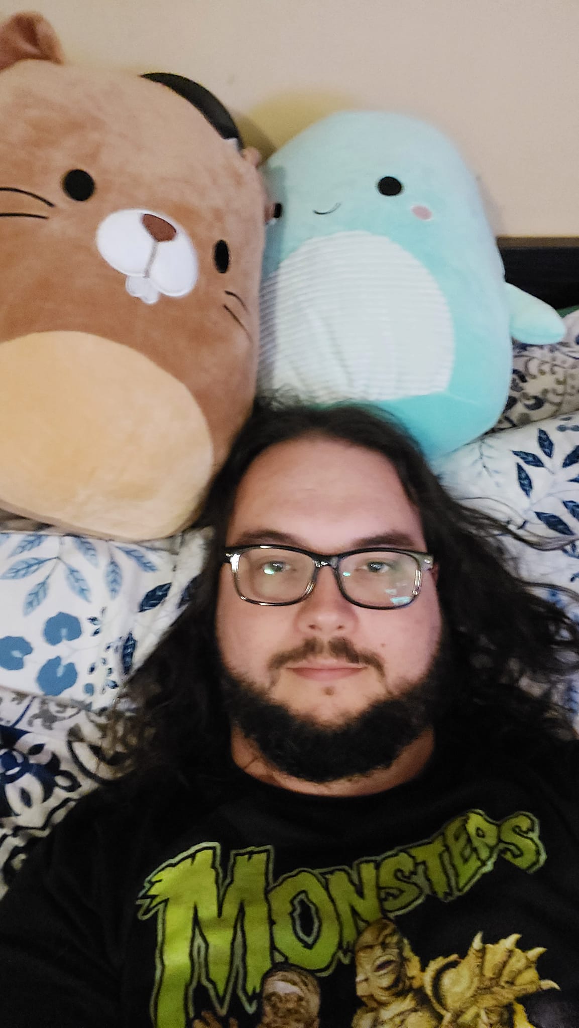 A man with long dark hair and a beard lies in bed with two Squishmallows, a beaver and a Nessie, photo 2